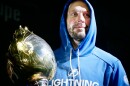 man with beard in blue hoodie holds large gold trophy 