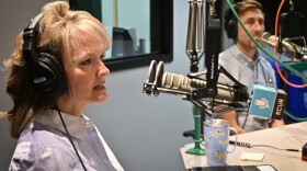 A woman, seated left, and a man sit inside a radio studio at microphones. The woman on the left is talking while the man is listening.