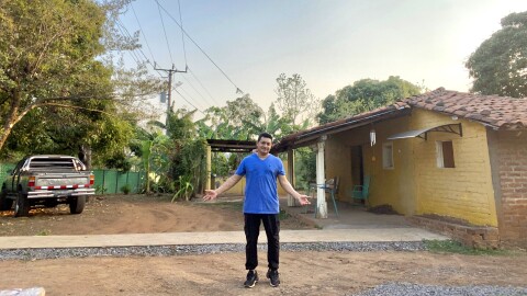 A man in a blue shirt stands in front of a house on a dirt road with his arms out