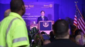 Craig Greenberg stands at a lectern in front of a blue background with an American flag nearby. A crowd of people, out of focus, watches him, including a person in a neon work vest who is standing to the left of the frame.