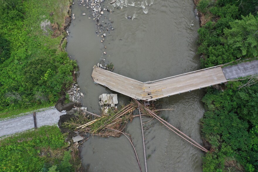  A wooden bridge over a river is snapped at an angle, and the gap is filled with trees and other debris