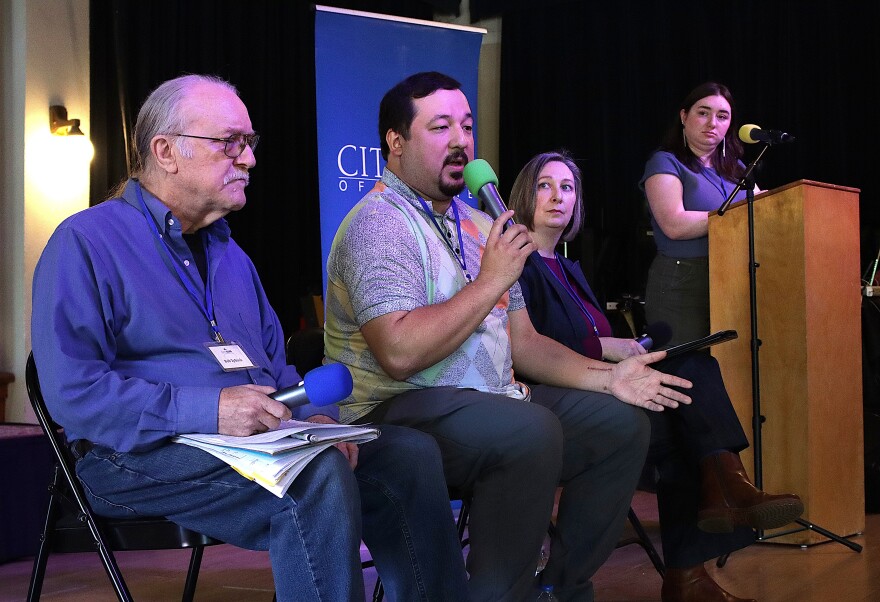  A group of people sit on a stage, one person speaks into a microphone, one person is at a podium.