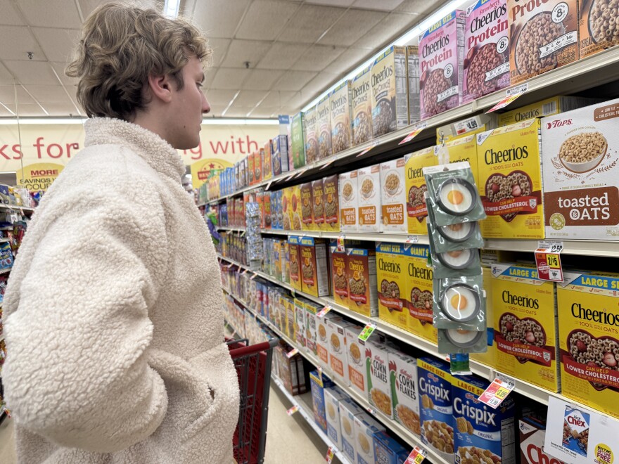 Reporter Boone Kilpatrick inspects price points on a grocery cereal aisle with bright colored boxes of Cheerios, Corn Flakes, and Chex Mix brands.