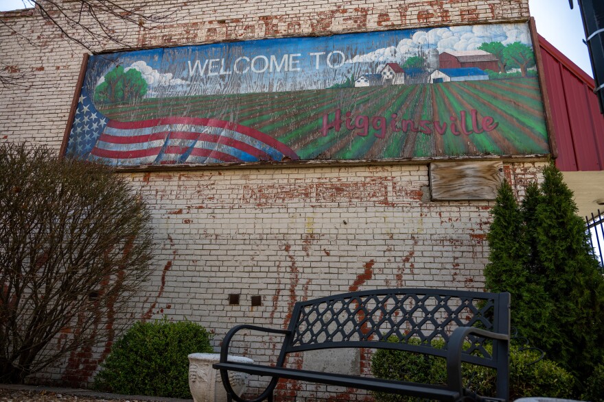 A tan brick wall with a mural on it shows an American flag on the left, and a field with some barns in the back. The words “Welcome to Higginsville” are on the mural. A bench and some bushes are in front of the wall.