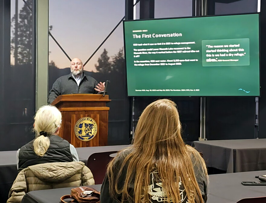 A speaker gestures behind a wooden podium at Oregon Tech beside a presentation slide about reconnecting Lower Klamath Lake and the Klamath River. Audience members sit in the foreground.