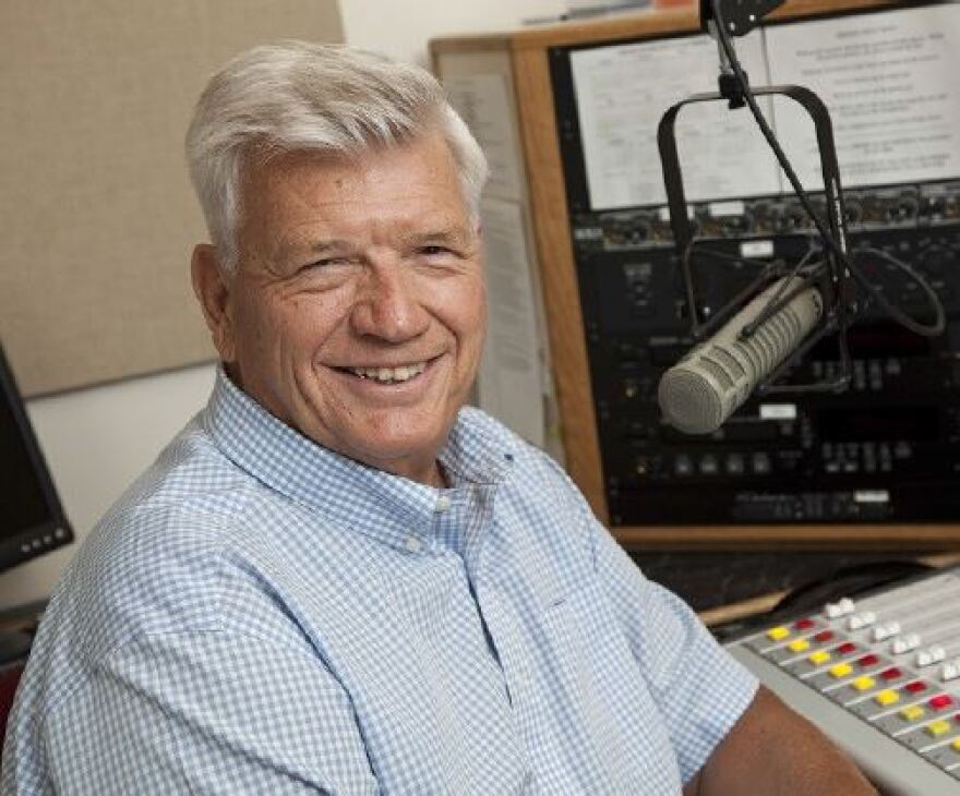 Bob Carroll sits in front of a microphone and mixing board inside a recording studio. 