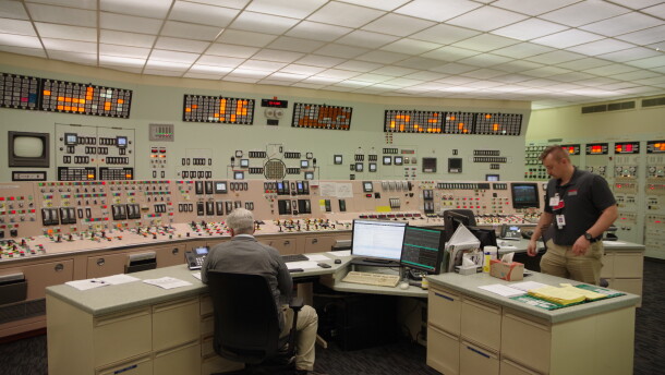 A large room has panels floor-to ceiling covered in buttons, levers and meters of all kinds. At the top of the panels are a series of orange lights. In the middle of the room, a man sits at a long desk in front of two computers. Another man stands near the desk. 