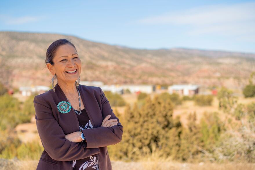 Woman, wearing a purple suit, stands with arms crossed in front of an arid, mountain landscape.