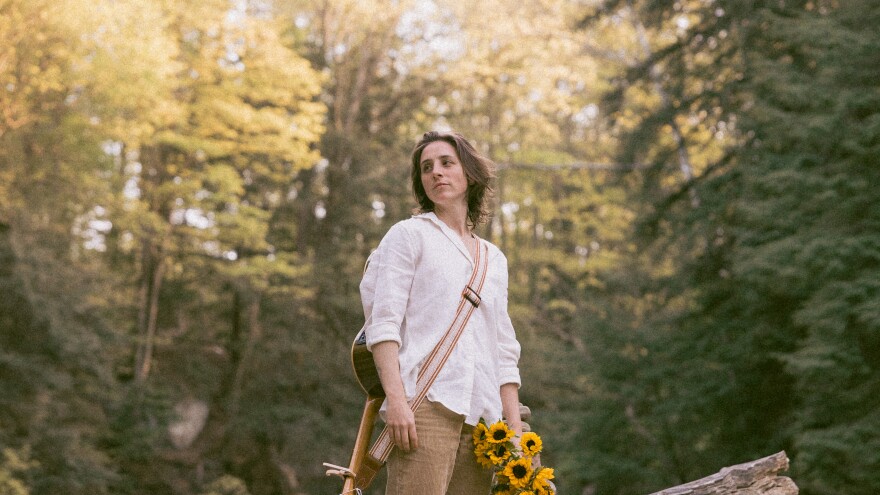 Tori Kurtz stands outside in nature with a guitar and sunflowers