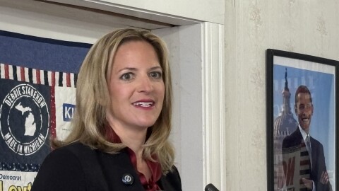 Michigan Secretary of State Jocelyn Benson stands at a podium with microphones, speaking indoors near a doorway. A campaign poster and a framed photo hang on the wall behind her. She wears a dark jacket over a light shirt and gestures with one hand while speaking.