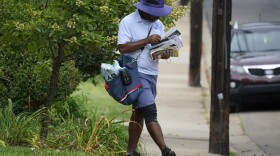 A US Postal Service carrier walks his route while carrying a bag full of mail and wearing a sunhat, shorts and short sleeved shirt, all in blue.