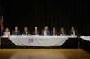 Seven men and a woman sit at along table. They're flanked on the stage by a table of a few journalists and a woman standing while speaking into a microphone. The ensemble looks a bit like "The Last Supper."