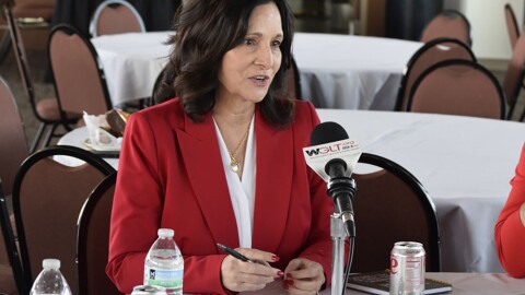 Woman in a red jacket and white shirt seated at a roundtable and speaking into a microphone 