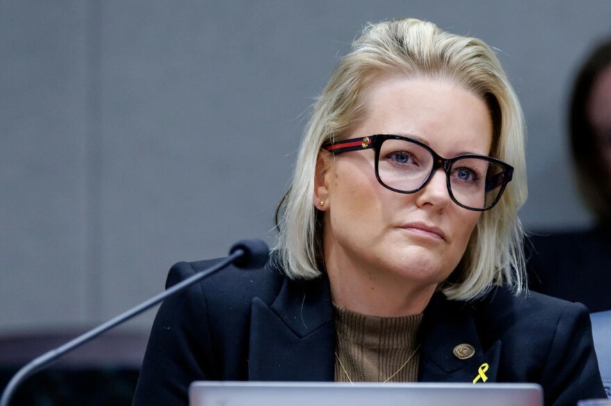 Blonde woman with glasses sits near a microphone. 