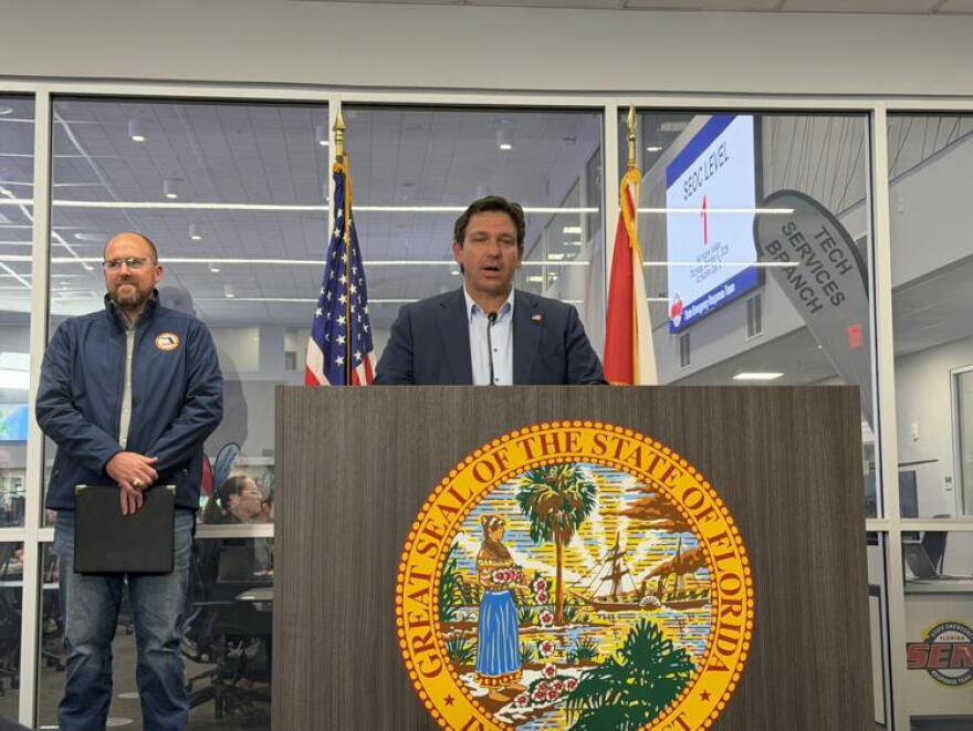 Man with brown hair and a dark suit jacket standing behind a podium with a yellow state seal, in front of a glass window and a man in a blue windbreaker to the left