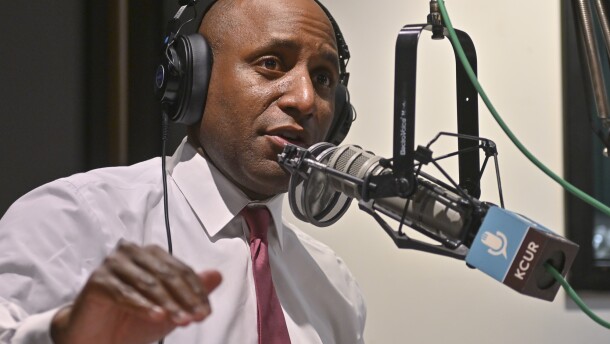 A man inside a radio studio sits behind a microphone. He is talking and gesturing with hie right hand.