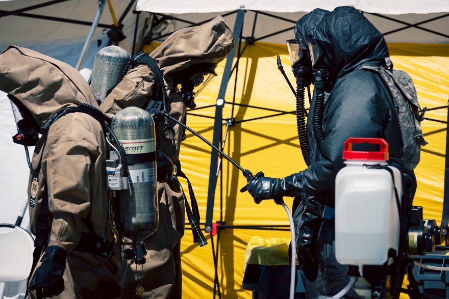 Two people in emergency response suits and masks walk past tents. 