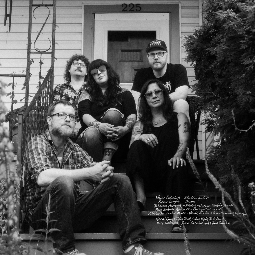 Group band photo of members of Chanteymen (from left to right): Christopher Lander Moore, Ethan Debelak, Johanna Andronis, Mary Antonia Andronis, and Ryan Lundin. They are sitting on the front steps of a house in a black-and-white photo.