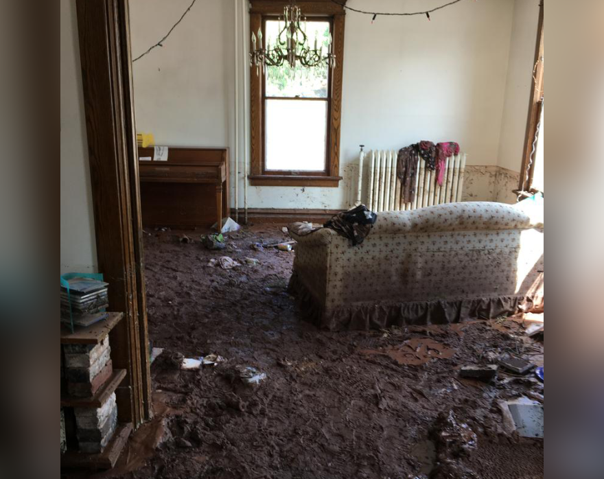 A living room, with a couch and a window, is shown with inches of mud piled on the carpet. 