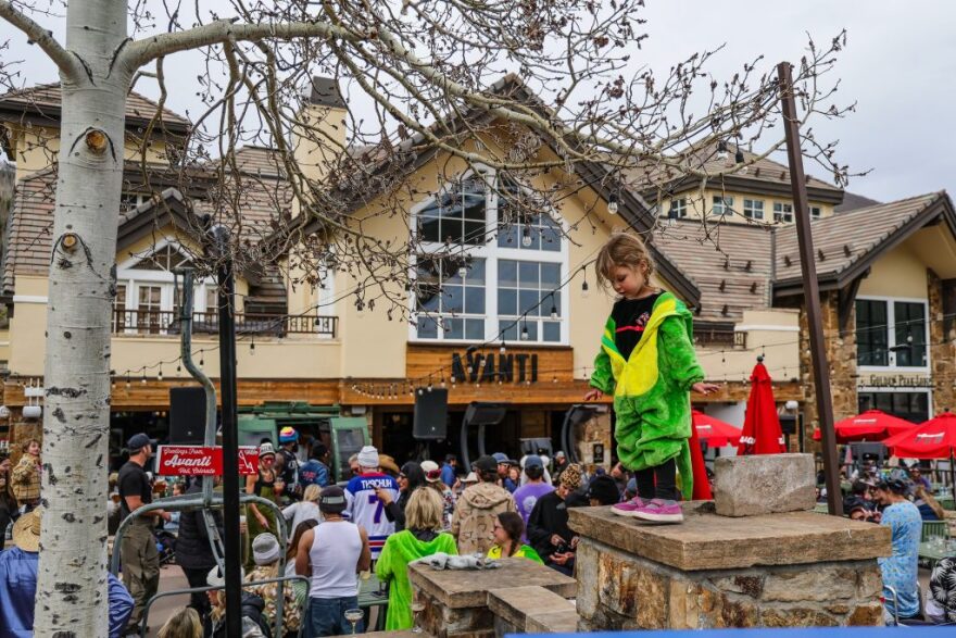 A large crowd stands in front of a building while a child in a dinosaur costume plays outside.