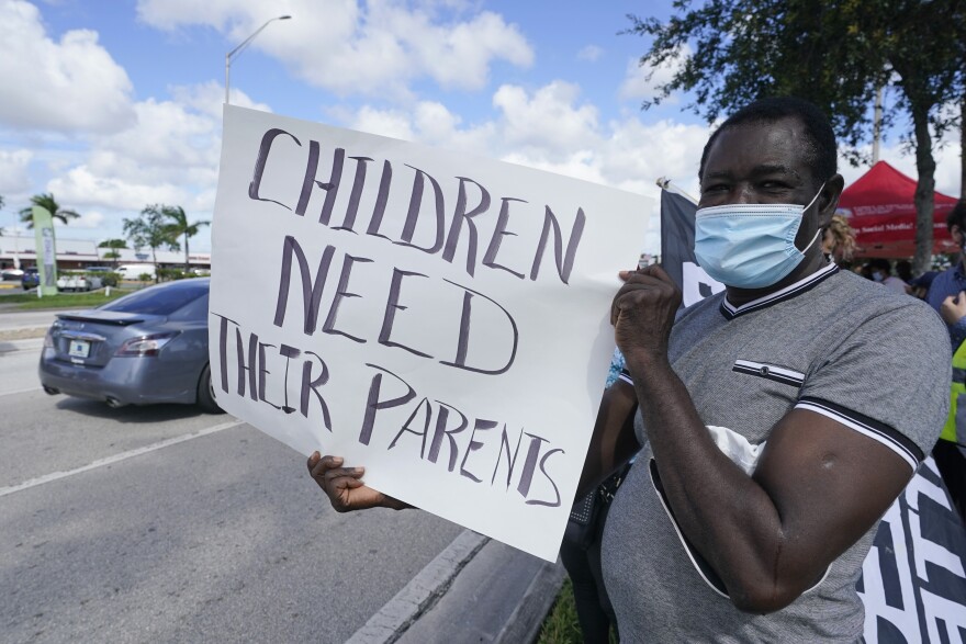 A man wearing a face mask holds a sign that says "CHILDREN NEED THEIR PARENTS" outside.