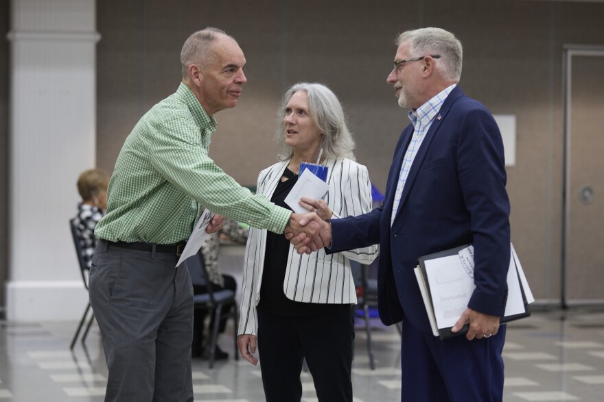 A man in gray pants and a green button down shirt shakes hands with a man in a navy suit as a shorter woman with gray hair and a white pinstripe blazer looks on.