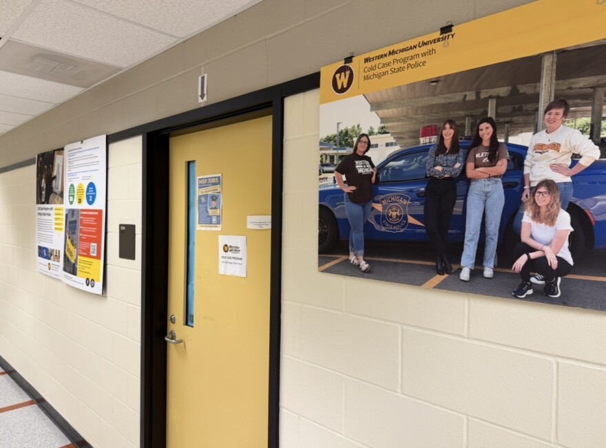 A wooden door is seen in a long white hallway. Next to the door, there are posters with information about the cold case program.