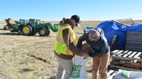 One person hold open a large white bag as another pours seeds from a paper bag into it.