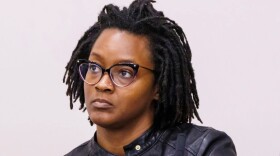Black woman with glasses, nose piercing and short dreads wearing a black leather jacket and looking off in the distance