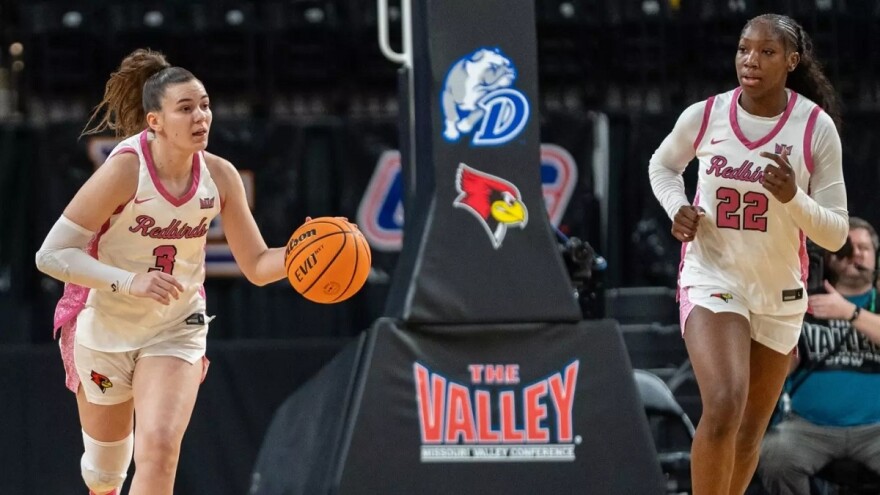Illinois State's Trista Fayta [left] dribbling a basketball and Doneelah Washington jogging alongside during a game with a basket support behind them featuring a logo that reads 'The Valley' and a redbird logo for Illinois State and a logo featuring a bulldog and the letter D for Drake 
