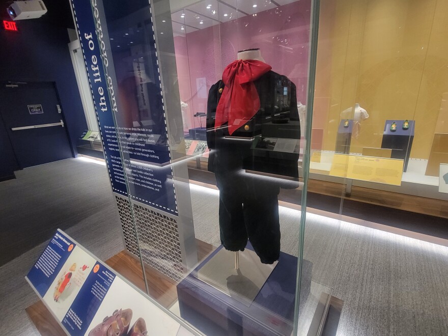 A child's outfit on display in the exhibit "The Life of Kids Clothes" at the Missouri History Museum. The outfit is of dark blue material, with gold-colored buttons and a large red ribbon. 