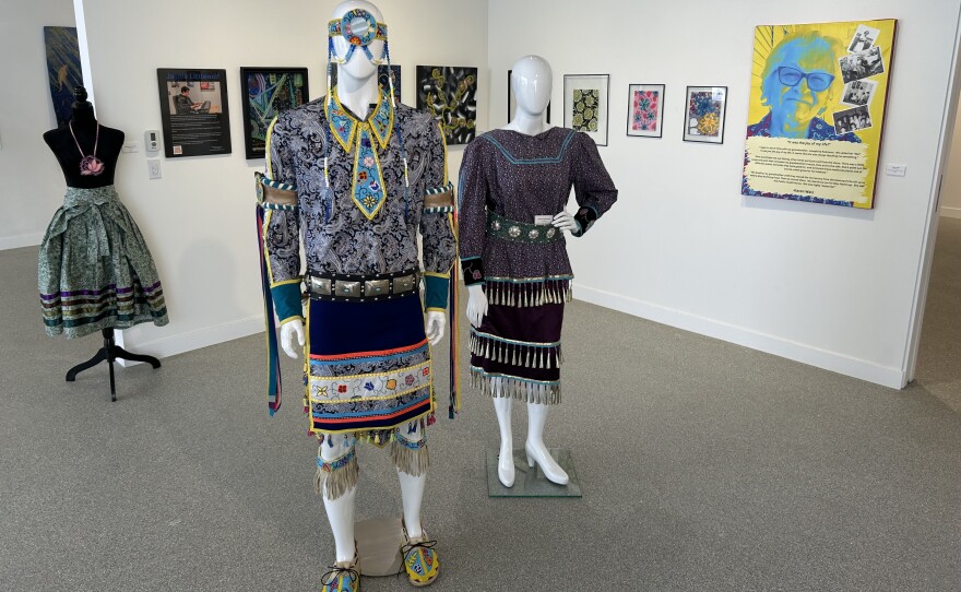 Two mannequins wearing Native American fashion and paintings in the background on a white wall.