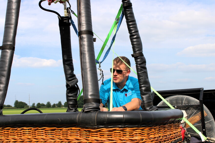 A man in sunglasses leans on the edge of a hot air balloon basket outdoors, with a field and blue sky in the background.