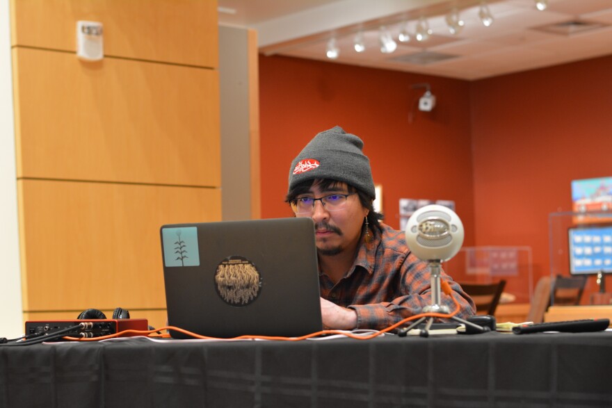 A young Native American man sits at this laptop. A ball-style microphone is on the table next to him.