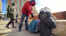 Two people wearing heavy coats and hoods approach a small collection of personal items leaning against a short concrete wall.. There is a rolling suitcase with two blue sleeping bags attached, a round, blue tent folded up and a small fabric bag lying nearby.