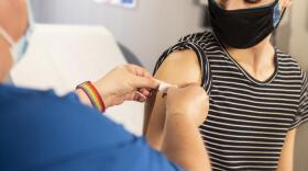 A nurse in blue scrubs puts a band-aid over the injection site for a young man in a black and white striped shirt.