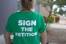 A woman, standing in front of tree, wears a green T-shirt that says "SIGN THE PETITION." She's only visible from behind.