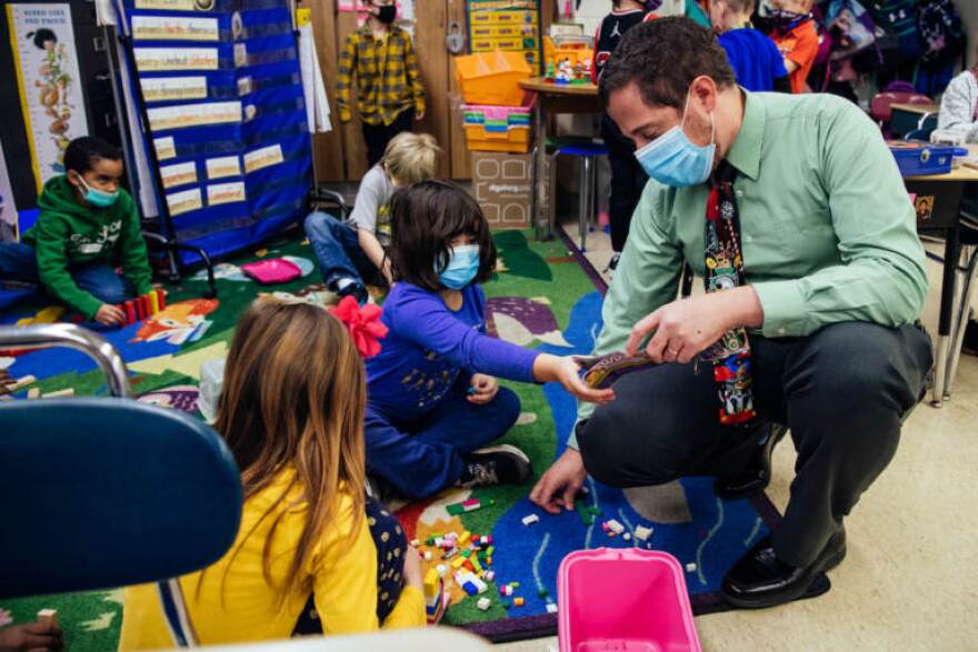 Principal Palazzo kneels beside students building with Legos so they can show him their progress during recess in a first grade classroom.