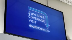 A tv screen mounted high on a white wall in a post office. An image with a plain, bright blue background fills the screen. In the center of the screen, in a mix of script and block fonts, are the words "If you lose coverage visit HealthCare.gov," with a thin blue arrow going from about "if" to the website url. A white outline of an arrow surrounds the blue arrow. 