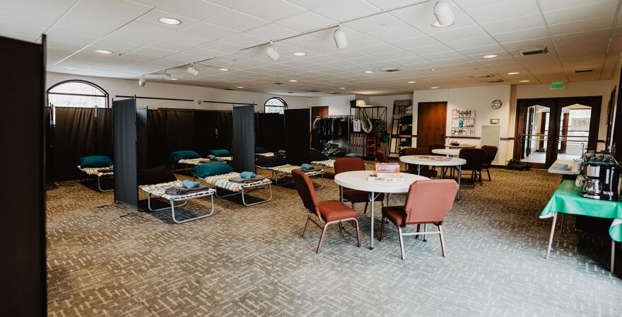 A room is filled with cots that are divided by curtains. There are several tables scattered around the room that have games on them.