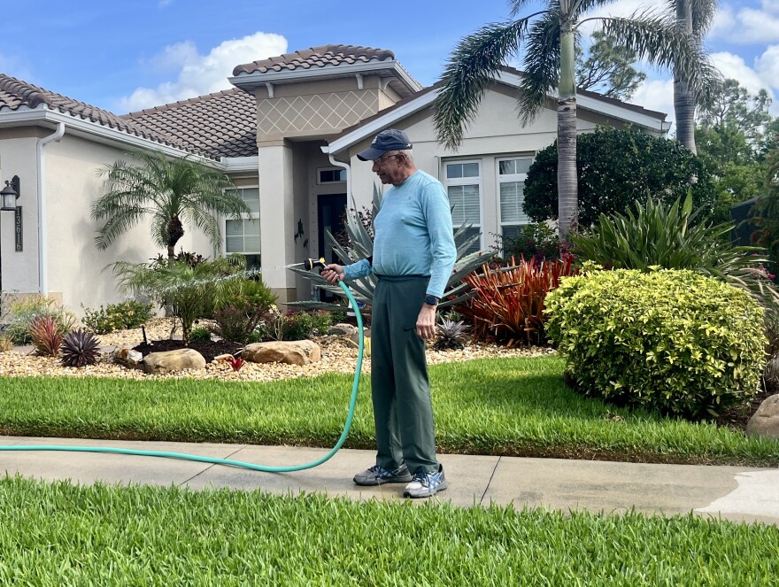 An older man in a blue shirt and cap watering a lawn in front of a house
