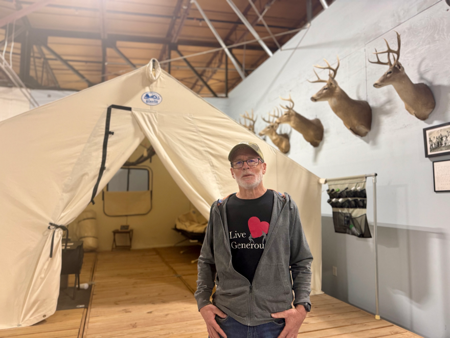 A man stands indoors beside a canopy tent. There are several deer heads mounted on the wall nearby.