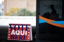 A sign says "Vote Aqui/Here" outside a building with someone inside at a voting machine.