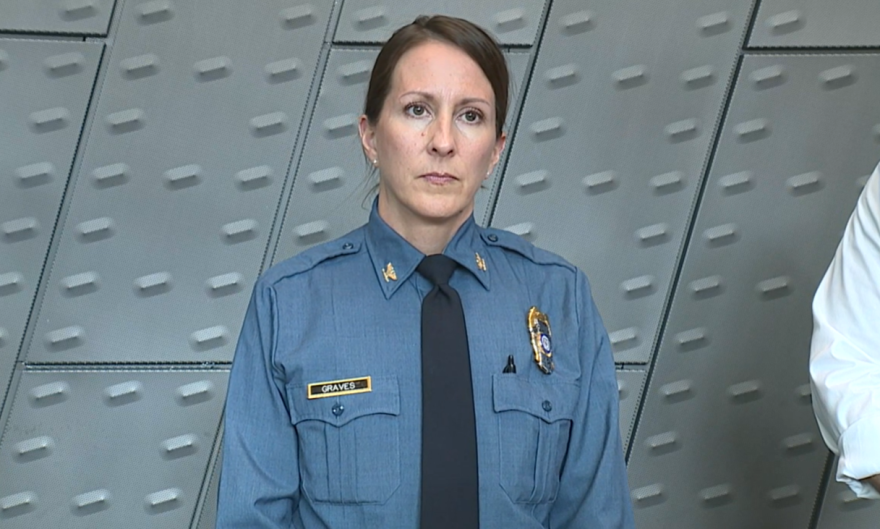 A woman in a blue police uniform looks straight ahead, in front of a grey background. 