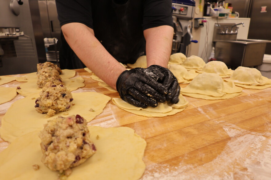 A baker uses two gloved hands to fold a large circle of dough in half, covering up the meat filling in the center.