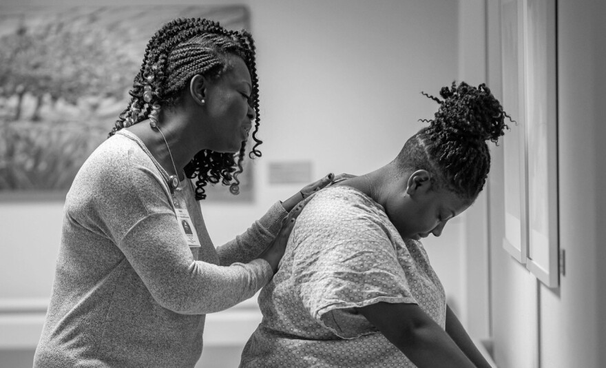 One woman standing behind and assisting another woman who's wearing a hospital gown