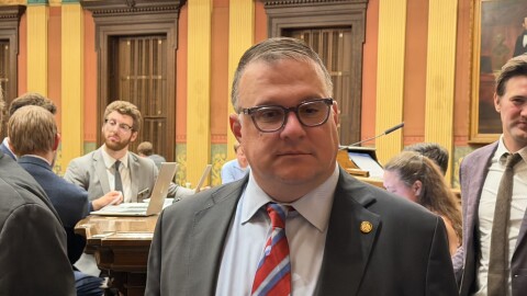 A man wearing a suit with a red, white, and blue striped tie stands in a yellow and orange room where people around him talk, walk, and work on laptops. He has gray hair, glasses, and is looking off from the direction of the camera.