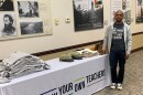 Man wearing a gray hoodie and dark shirt that reads "Grow Your Own Teachers" standing next to a table with a sign that also reads "Grow Your Own Teachers" while several hoodies, hats and tote bags are stacked on the table in a conference room