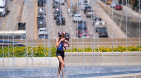A young girl with brown hair in shorts and a tanktop holding a doll walks through a fountain that overlooks a highway.