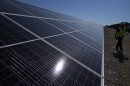 A close up of a ground mounted solar panel. A construction worker in a hard hat and a yellow safety vest walks past the row of panels in the background. 
